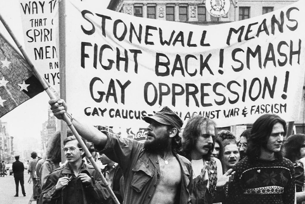 A scene from the Stonewall Riots. Men march down the street holding banners and signs. One says "Stonewall means fight back! Smash gay oppression! Gay caucus march against war & facism."